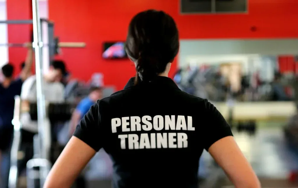 Trenerka personalna w koszulce z napisem Personal Trainer obserwuje salę siłowni